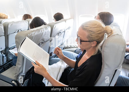 Woman reading magazine on airplane during flight. Female traveler reading seated in passanger cabin. Stock Photo