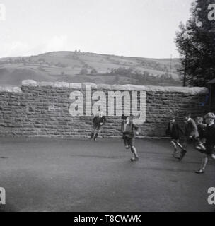 1950s, historical, young schoolboys playing football outside with a small ball in a stone walled school playground, with the rolling hills of the countryside in the distance, England, UK. Stock Photo