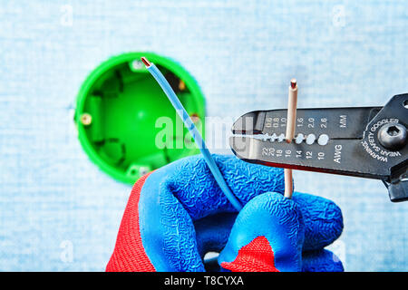 Electrician in protective gloves strips off the wire with wire stripper cutter during installing round electrical box. Stock Photo