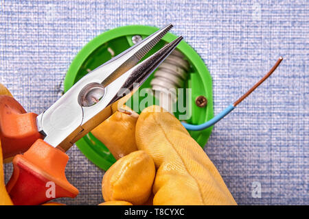 Electrician in protective gloves is cutting the ends of the wires of round outlet box with long nose pliers, electrical installation. Stock Photo