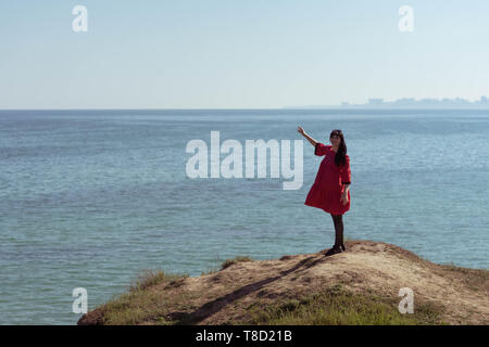 Beautiful brunette woman in red dress posing against the backdrop of the sea on a sunny spring day. Stock Photo
