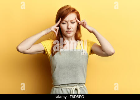 ginger girl keeps fingers on temples, tries to remember information, can find quick answer in mind, tries concentated on thougths, garderner, housewife having headache. isolated yellow background Stock Photo
