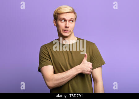 happy man shows thumbs up sign, expresses agreement, cheers his friend up, looks aside. isolated over violet background, studio shot. Stock Photo