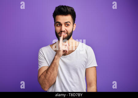cunning man wearing white t-shirt holding finger at his lips, asking to keep silent and not to make noise.studio shot. Shhh. stop talking, gossip concept. Stock Photo
