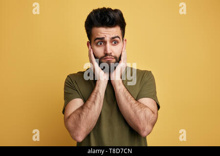 scared handsome man hears negative news, keeps palms on cheeks, expresses amazement. close up photo. isolated yellow background.emotion concept Stock Photo