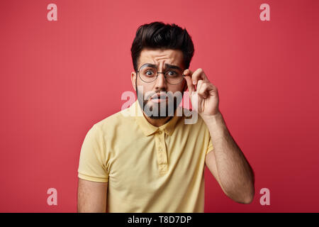 funny man with bad eyesight trying to see written on poster, board.expresses surprise , close up photo. isolated pink background.man in yellow T-shirt touching his eyeglasses and looking at camera Stock Photo