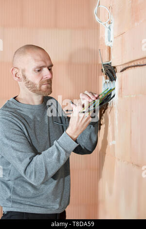 Electrician at work installing sockets in unfinished house built of clay block bricks. Worker wiring cables inside house under construction. Stock Photo