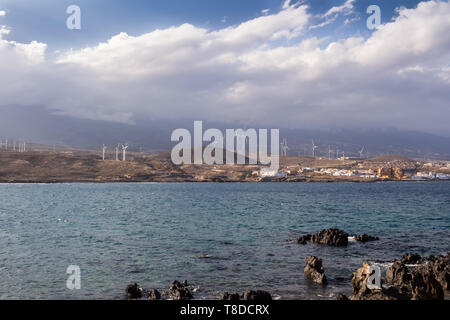 View from the coast on the Atlantic ocean waves. Wind electricity power stations, city and a mountain in the background. Punta de Abona, Tenerife Stock Photo