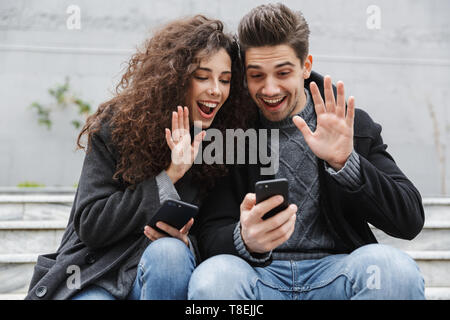 Image of cheerful couple man and woman 20s in warm clothes using smartphones together while sitting on stairs outdoor Stock Photo