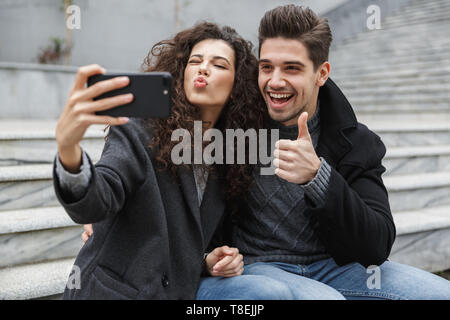 Image of excited couple man and woman 20s in warm clothes taking selfie photo on cell phone while sitting on stairs outdoor Stock Photo
