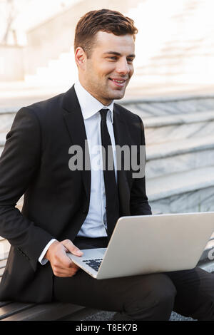 Photo of handsome young businessman in formal clothes outdoors at the street using laptop computer. Stock Photo