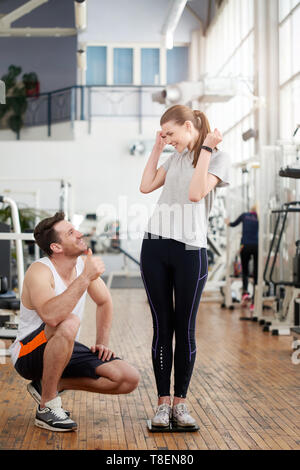 Fitness trainer showing thumb up to female client. Young excited woman celebrating her weight loss standing on scale at gym. Fitness achievements with Stock Photo