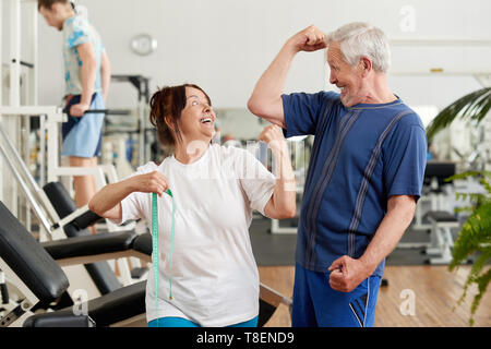 Cheerful senior couple showing their biceps at the gym. Joyful senior woman with measuring tape at fitness club. People, sport, leisure and healthy li Stock Photo