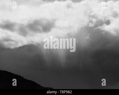Shades of grey, Wispy clouds and beams of sun light streaming and breaking through the fluffy cumulus clouds above pointing towards the ground Stock Photo