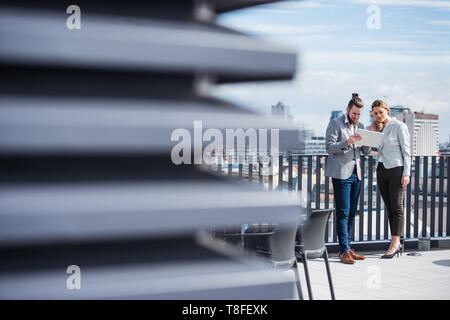 Two young business people with tablet standing on a terrace outside office, working.