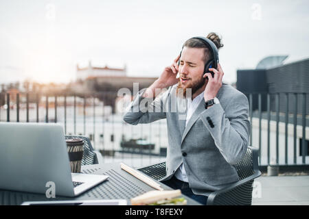A young businessman with headphones sitting on a terrace, having break. Stock Photo