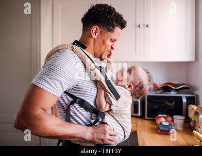 A father with small toddler son in carrier in kitchen indoors at home. Stock Photo