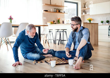 An adult hipster son and senior father sitting on floor indoors at home, playing chess. Stock Photo