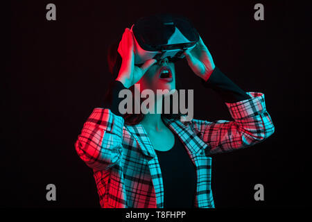 Side view portrait of pretty teenage girl wearing virtual reallity glasses with cyan and red color gels in studio over black background. Teenage girl using modern techonlogy. Cheerful young lady using vr headset. Stock Photo
