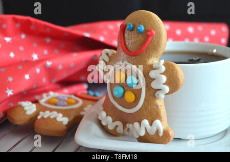 White cup of tea with a gingerbread man cookie, another gingerbread man with beautiful colorful sugar icing close by, red starred christmas fabric, wh Stock Photo