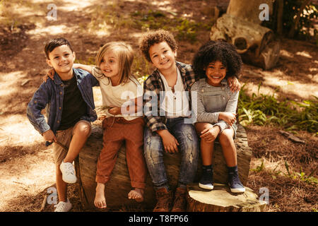 Group of four kids sitting on a wooden log outdoors. Multi-ethnic group of kids playing together in a forest. Stock Photo