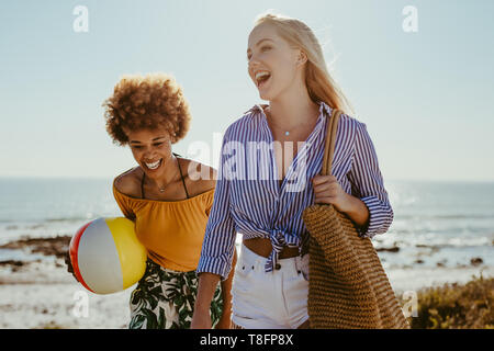 Two young women walking with a ball and bag along the beach. Female friends going on picnic at beach.