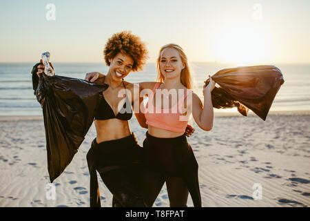 Two young female volunteers with garbage bag. Two surfers standing on the beach with plastic garbage bags with sunset in background. Stock Photo