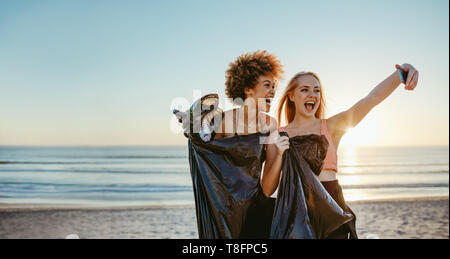 Female volunteers with garbage bag taking selfie on the beach after doing trash tag challenge. Stock Photo