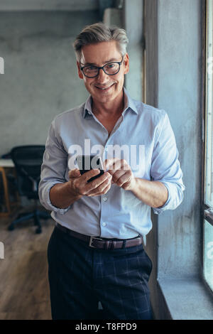 Happy mature businessman standing by a window with his mobile phone. Male entrepreneur at his office. Stock Photo