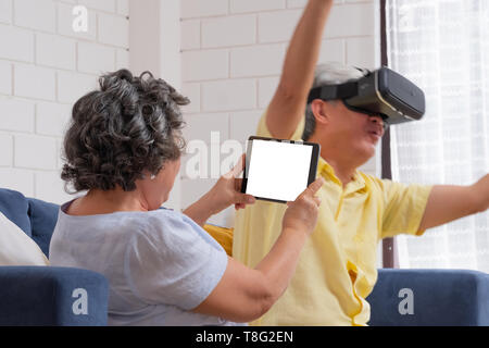 Asian senior couple play virtual realtiy glasses headset and tablet watching vr video and  have fun together on sofa  in living room at home.senior wi Stock Photo