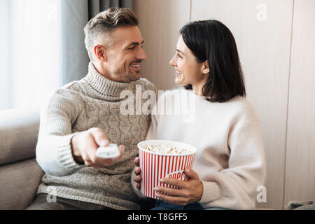 Image of a loving couple family sitting on sofa indoors on sofa watch tv with remote control eat popcorn. Stock Photo