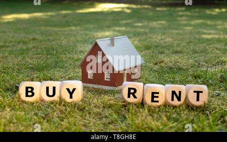 Concept for buying or renting a house. A miniature house placed on green meadow. Dice form the words 'BUY' and 'RENT'. Stock Photo