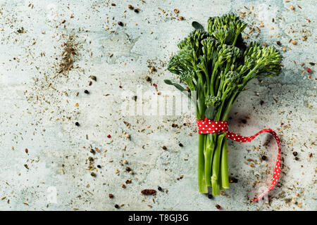 high angle view of a bunch of broccolini, tied with a red ribbon patterned with white dots, placed on a white stone surface, with some blank space on  Stock Photo