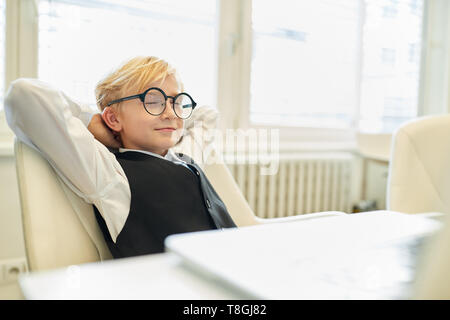 Relaxed child sits as a boss in the office with eyes closed while dreaming Stock Photo