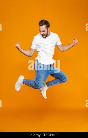 Full length photo of smiling guy in t-shirt and jeans jumping and having fun isolated over yellow background Stock Photo