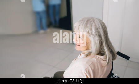 Smiling senior woman in wheelchair sitting at the hospital corridor. Go home after discharge from hospital Stock Photo