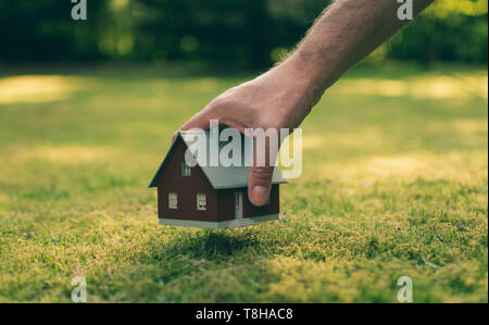 Concept of selling a house. A hand is holding a model house above green meadow. Stock Photo