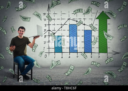 Young man sitting on chair holding laptop in one hand and mobile phone in another, celebrate business success, showing a positive increasing graph of  Stock Photo