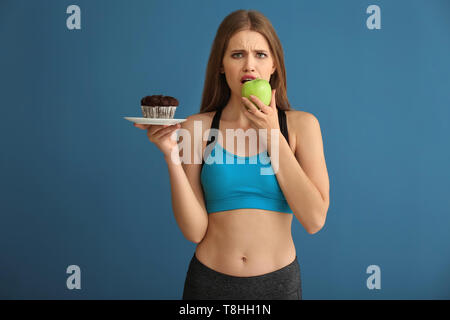 Young woman in sportswear choosing between cake and apple on color background Stock Photo