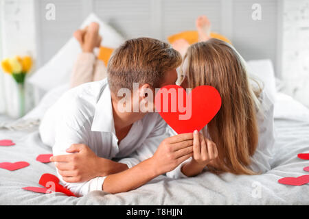 Happy young couple hiding faces behind red heart while lying on bed at home Stock Photo