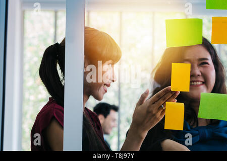 Female office managers smiling while looking at note stickers on glass wall in modern office Stock Photo