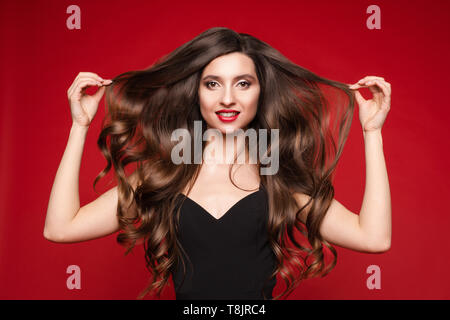 Studio portrait of stunning young caucasian woman with gorgeous long brunette hair in black with red lips. Isolate on red. She is showing beautiful, s Stock Photo