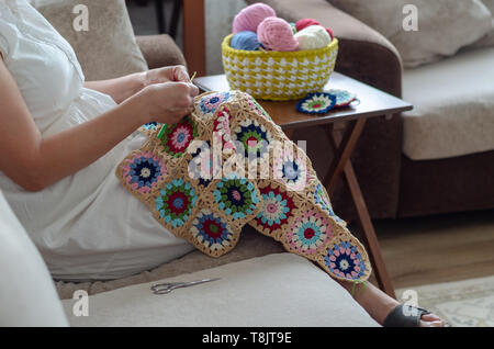 There are multi colored yarns in a basket next to the woman.On the background, the woman is crocheting  baby blanket with multi colored yarns Stock Photo