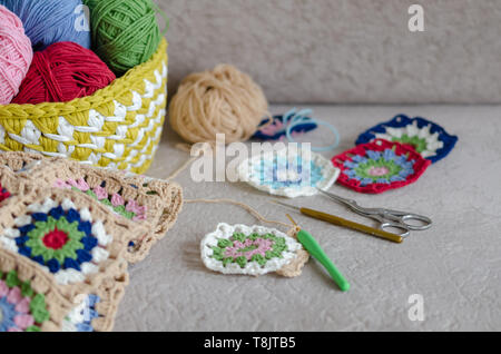 Multi Colored plaid squares of crochet on a cream colored seat background. Green, pink,cream, blue,red color yarns,hookeds, scissors are  on the sofa. Stock Photo