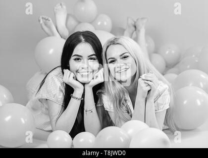 Gossip concept. Blonde and brunette on smiling faces have fun at domestic bedroom party. Girls lay on belly near balloons, pink background. Sisters or best friends in pajamas at girlish pajamas party. Stock Photo