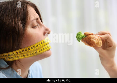 Offering of tasty croissant sandwich to young woman with measuring tape around her mouth on light background. Diet concept Stock Photo