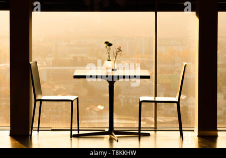Two minimalist chairs and a table set for a romantic dinner in a luxury restaurant with a panoramic city view during golden hour sunset Stock Photo