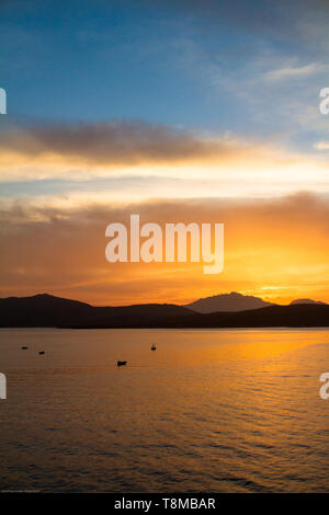 Sunrise on the Sardinian sea coast with intense orange color seen from the sea with five fishing boats on flat water Stock Photo