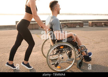 Sporty woman helping young man in wheelchair outdoors Stock Photo