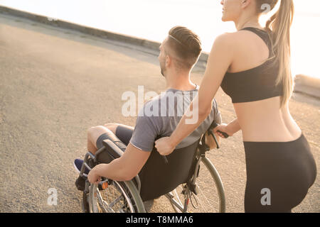 Sporty woman helping young man in wheelchair outdoors Stock Photo